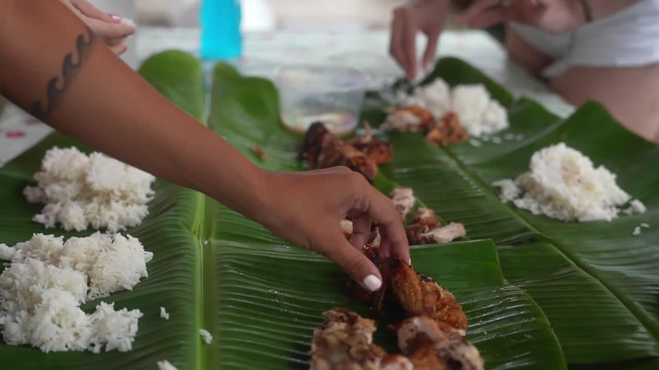 Close-up handheld shot captures hands placing grilled chicken and rice onto banana leaves in a traditional Philippine boodle fight, highlighting communal dining and cultural warmth