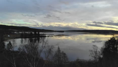 Drone-rising-over-trees-by-a-lake-in-Sweden-at-sunset