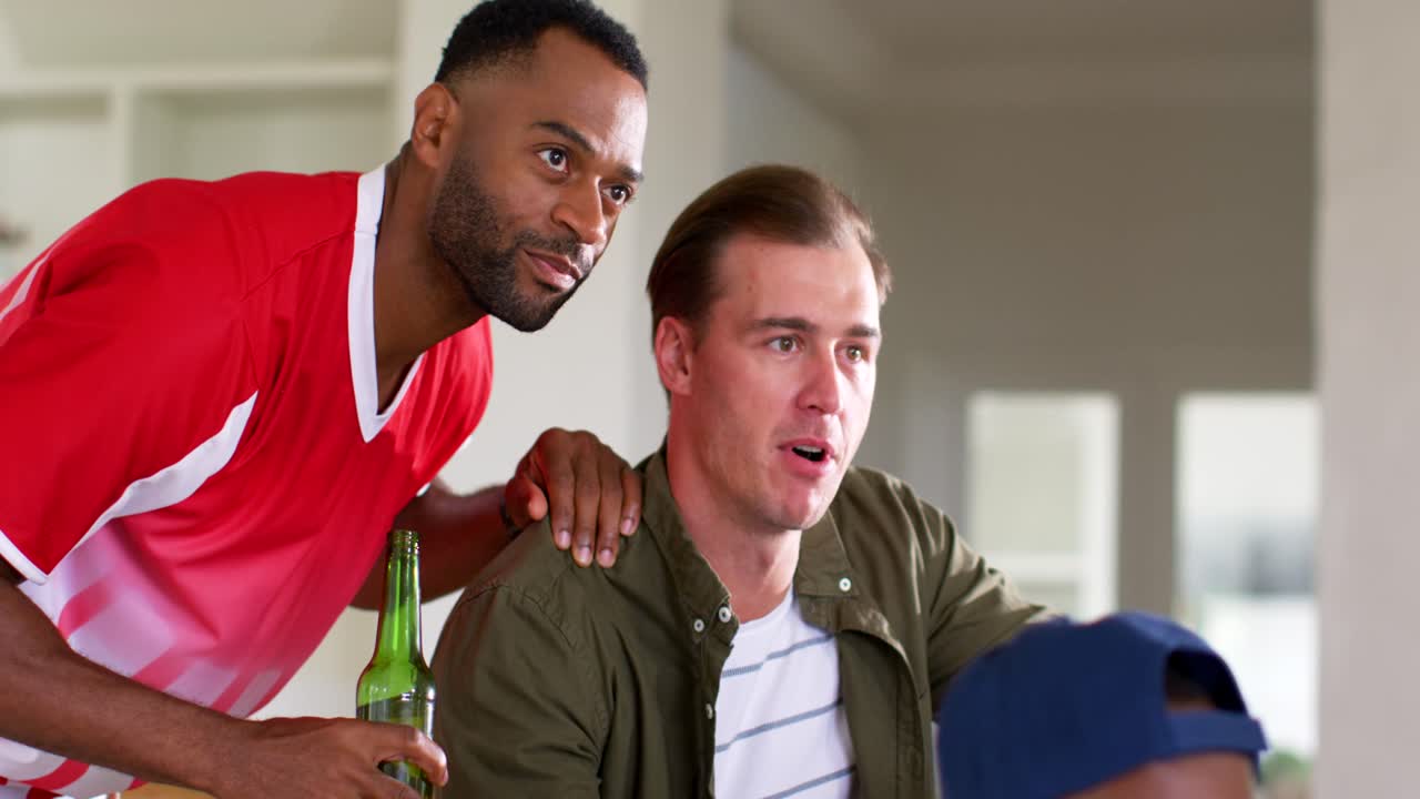 Diverse male friends at home red jersey leaning watching game event holding remote sipping beer