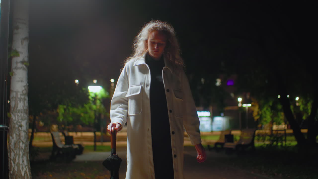 Elegant girl walks confidently through open outdoor space at night holding umbrella, illuminated by gentle streetlights surrounded by park benches, trees, and soft background lighting