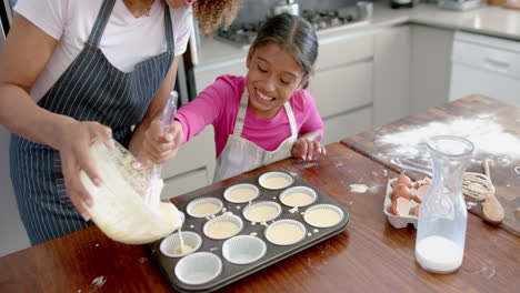 happy biracial mother and daughter pouring cake mix into forms in kitchen, copy space, slow motion