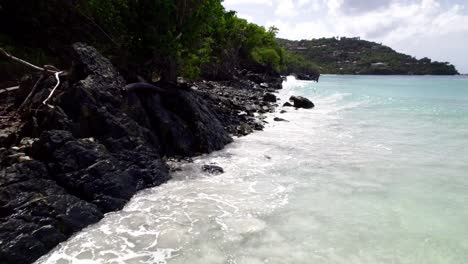 Stunning-Aerial-Wide-shot-of-water-crashing-on-rocks-and-the-beach-shore-sand-buildings-on-hill-in-background-blue-sky-white-clouds-turquoise-water-relaxation-vacation-tourism