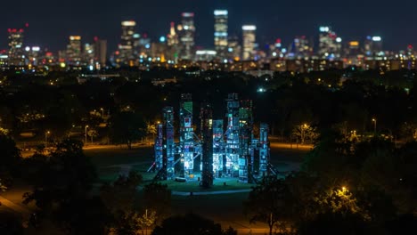 illuminated artistic installation against a glowing city skyline at night, showcasing modern architecture and vibrant lighting effects in a lush urban park
