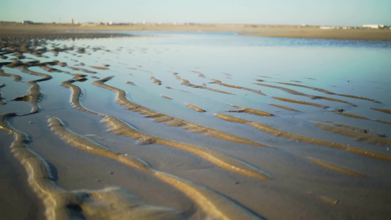 Shifting low camera revealing wet-sand ripples and shallow tidal pool on tidal flat, showing houses
