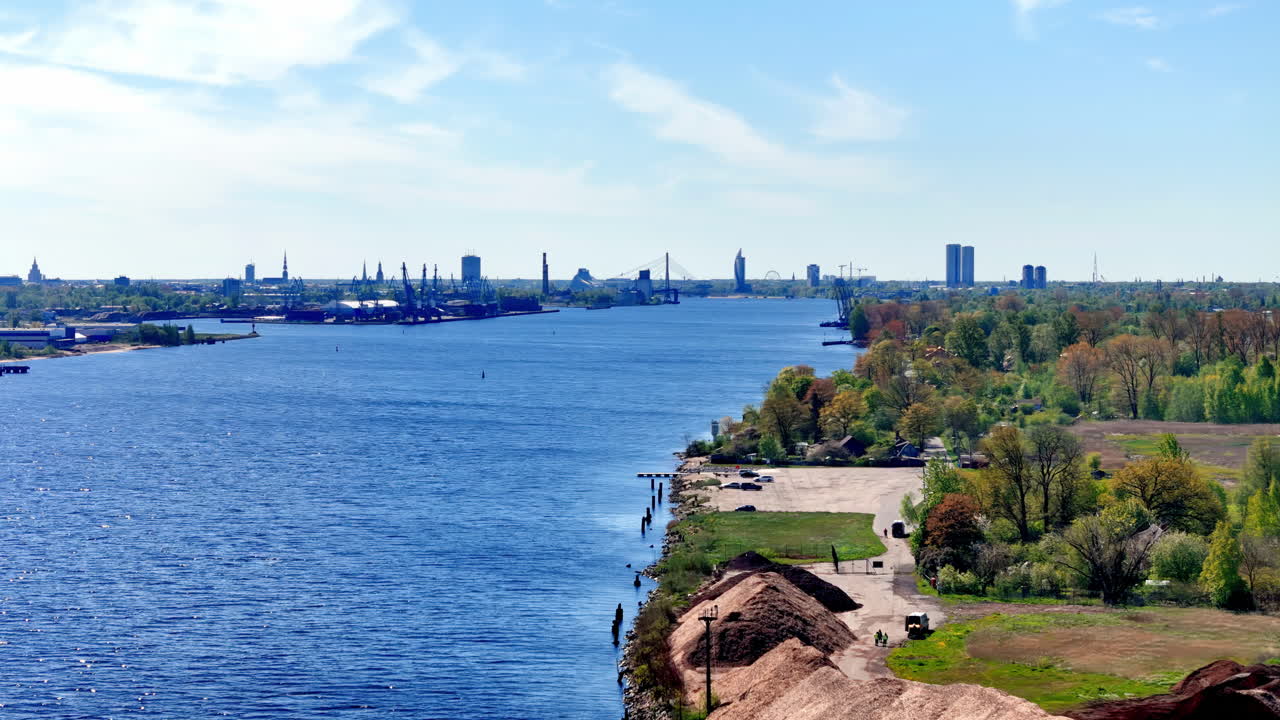 aerial shot on a bright, clear day in late spring early summer, capturing a wide blue river flowing towards a distant city skyline under a partly cloudy sky. In the foreground, a lush green riverbank