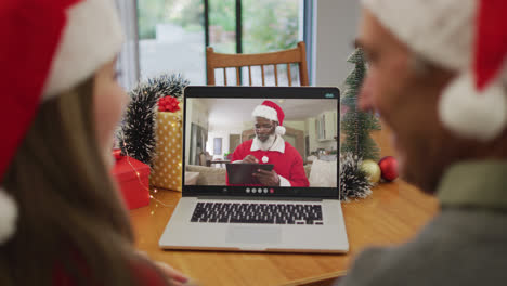 Caucasian-couple-on-laptop-video-call-with-santa-claus-at-christmas-time