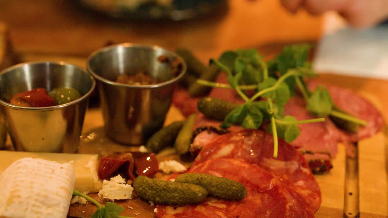 Hand uses fork to pick up pickle from charcuterie board under warm indoor lighting