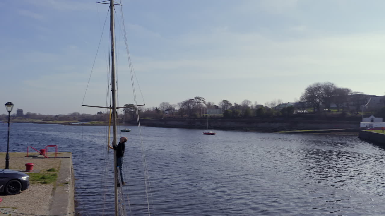 Drone wide orbit of man climbing mast rigging and preparing sunken sailboat, reflection shimmering on quiet waterline, Kinvara harbour, Galway Ireland