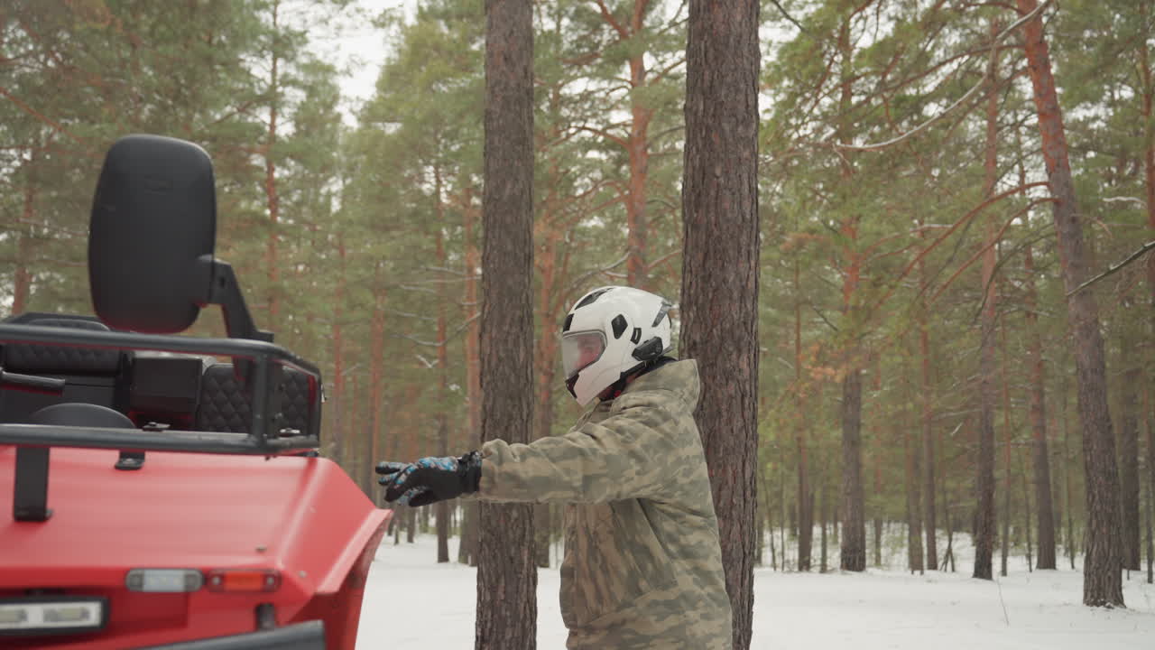 Pilota nella pineta innevata prepara il quad, indossando casco e guanti, si avvicina a un quad rosso parcheggiato tra pini alti, giacca mimetica, atmosfera fredda e silenziosa, neve soffice sotto i piedi, movimenti cauti e concentrati.