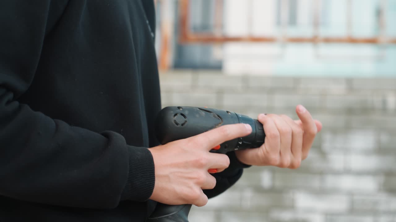 Mani in primo piano che tengono un trapano a batteria, preparando la punta all'aperto contro un muro di cemento, regolando l'impugnatura e il grilletto con movimenti costanti, manica di maglione nero visibile, preparazione calma e concentrata per un progetto di riparazione