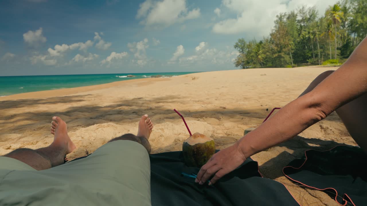 Primo piano delle gambe di una coppia che si rilassa su una spiaggia tropicale con noci di cocco e oggetti arrotolati appoggiati su un asciugamano. Onde assolate, sabbia e palme creano un'atmosfera vacanziera pacifica e rilassata.