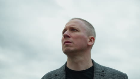 a close-up view of an aged white man wearing a grey suit and a black polo shirt, focusing his attention on the right hand side, with an overcast sky in view