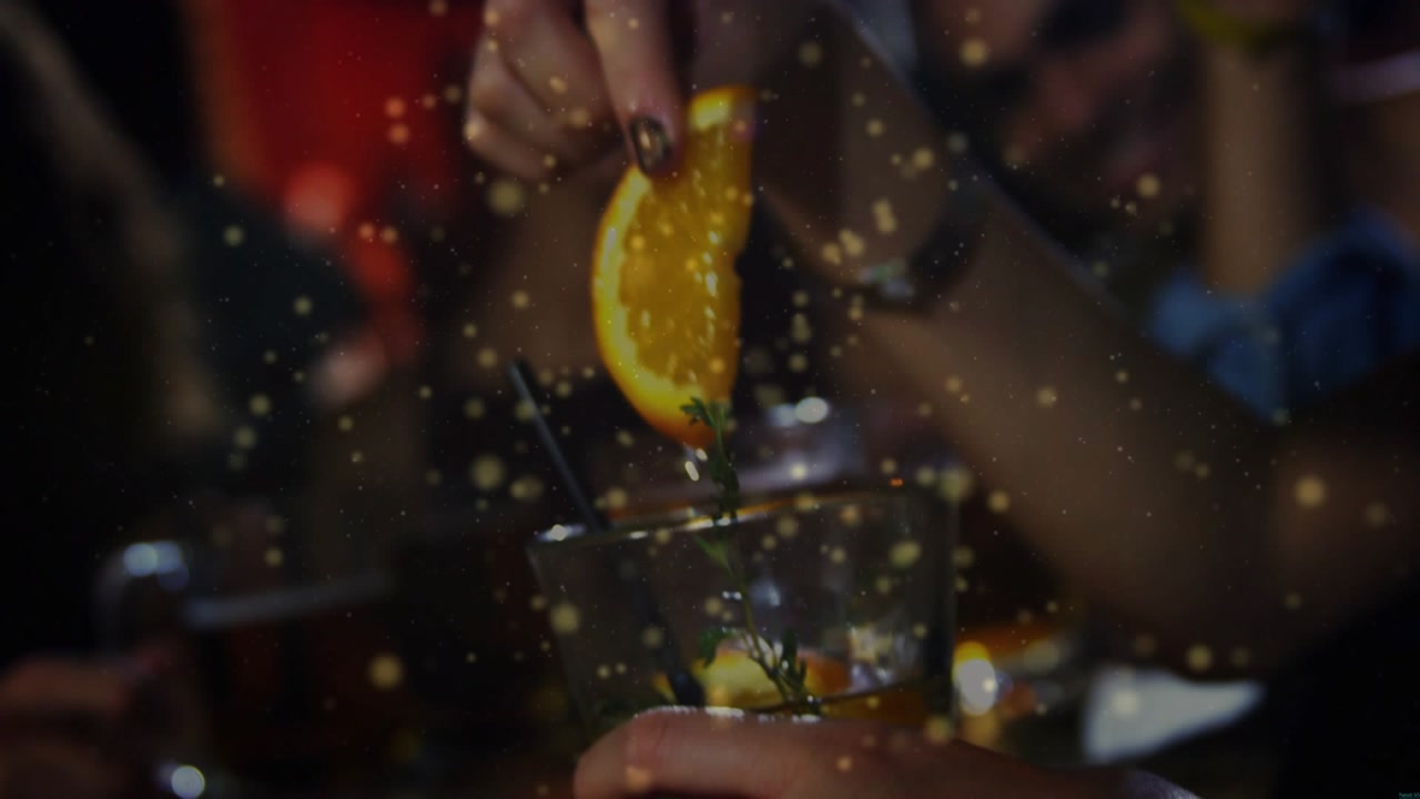 Man holding lowball at bar, woman lowering, pressing orange wedge into drink for aroma, golden glow