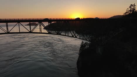 Wide-aerial-shot-of-car-silhouettes-driving-across-Deception-Pass-Bridge-at-sunset