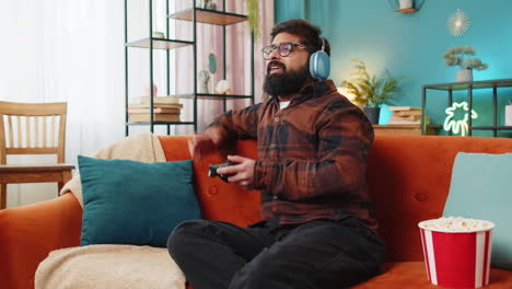 joven indio usando un controlador de joystick jugando un juego de consola de video juego en línea en el sofá de casa