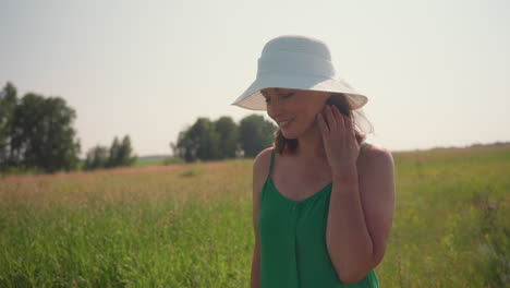 Smiling woman in green dress and wide white hat walks peacefully through grassy field on sunny day, gently touching hair while enjoying warm breeze and nature under clear sky in serene countryside