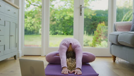 Young-Woman-Sitting-On-Mat-At-Home-With-Laptop-Doing-Online-Yoga-Class