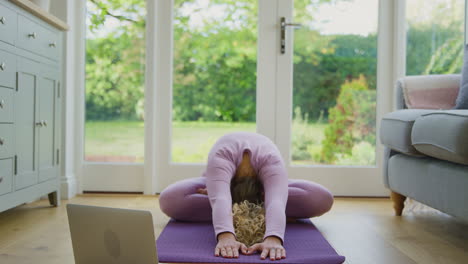 young woman sitting on mat at home with laptop doing online yoga class