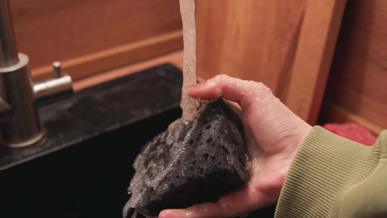 Close-up of hand wearing green sleeve holding dark porous sponge under steady tap stream while lathering and squeezing to create foam and droplets; warm wooden countertop and stainless steel sink in