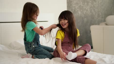 Lovely-little-cute-girl-combing-the-hair-of-her-older-sister-and-while-sitting-on-the-bed-at-home