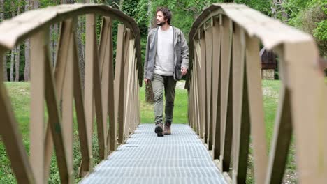 young man in his 20s wearing neutral clothing walking over a small footbridge towards the camera on an outdoors hiking trail in catalonia