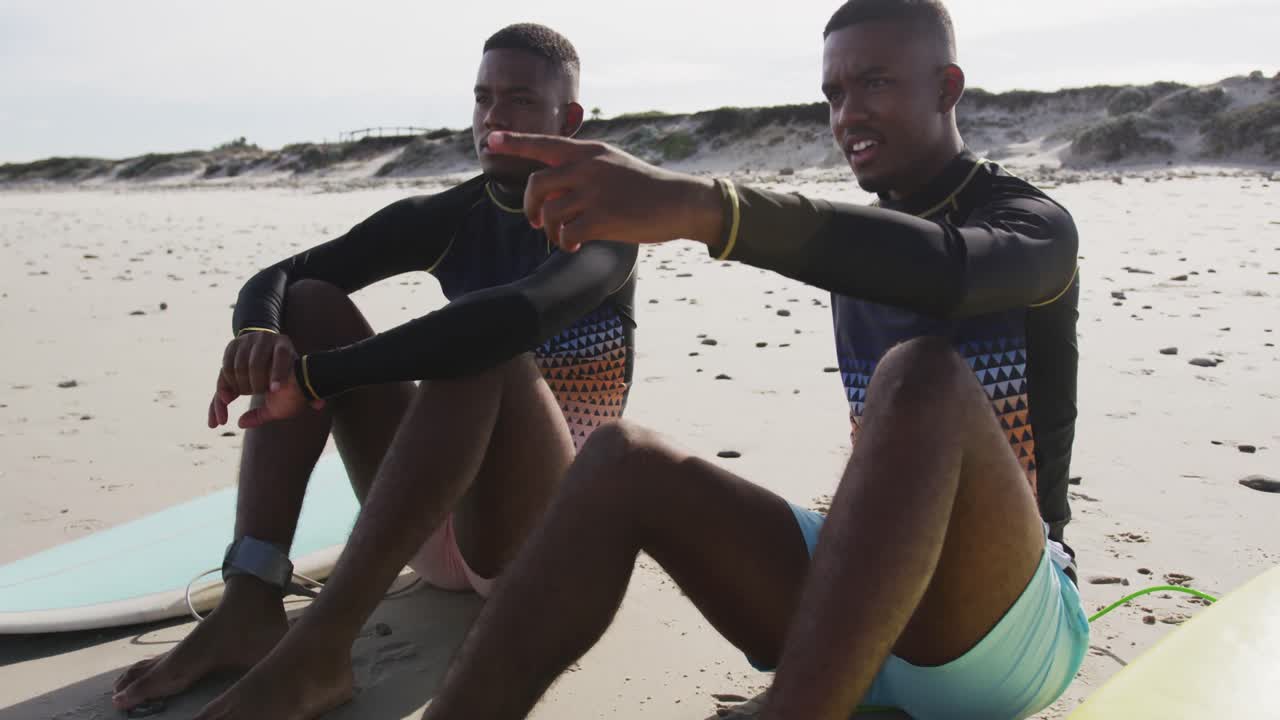 felices hermanos gemelos adolescentes afroamericanos sentados junto a las tablas de surf en una playa hablando