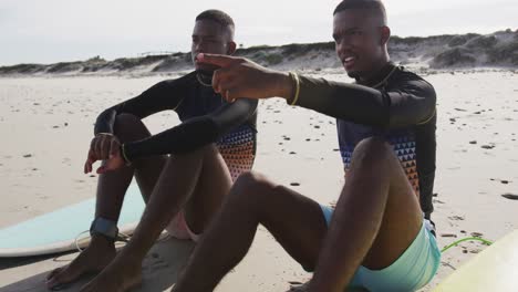 happy african american teenage twin brothers sitting by surfboards on a beach talking