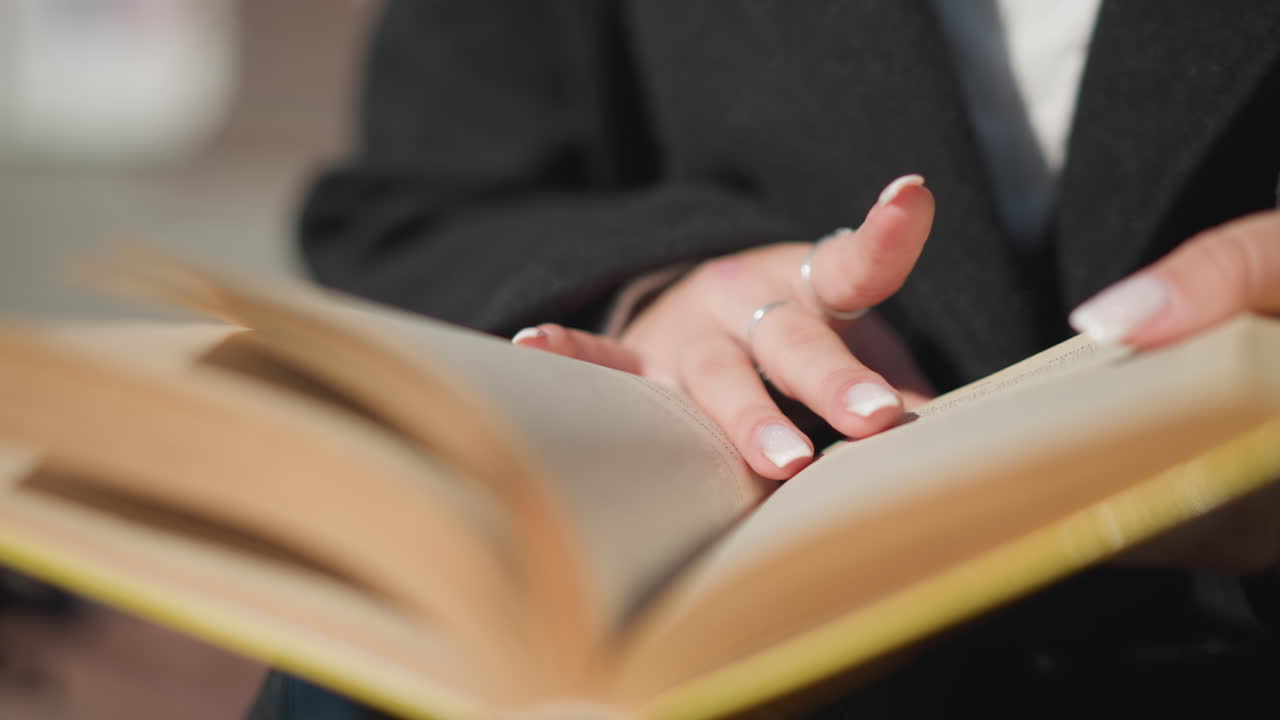 책 페이지를 뒤집는 손의 클로즈업, 반지로 장식된 은 손, 검은 옷을 입고, 배경은 보케 효과로 흐릿합니다.