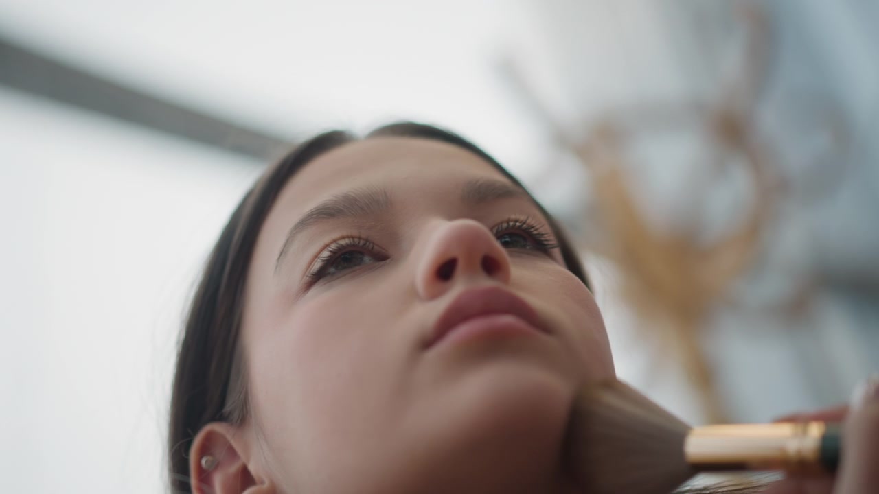 Low-angle close-up showing young woman during makeup brush application on cheek and jawline, with soft indoor light, visible skin compression, calm expression, beauty routine atmosphere