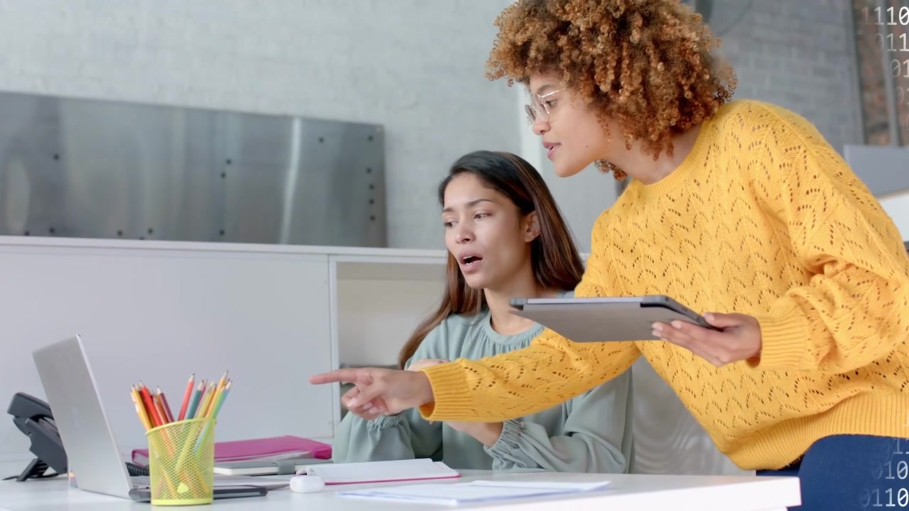 Seated woman gesturing peer leaning with tablet pointing at laptop reviewing IT with binary overlay
