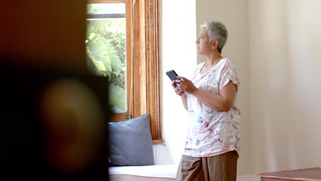 Mujer-Birracial-Mayor-Pensativa-Usando-Un-Teléfono-Inteligente-En-La-Ventana-De-Casa,-Cámara-Lenta