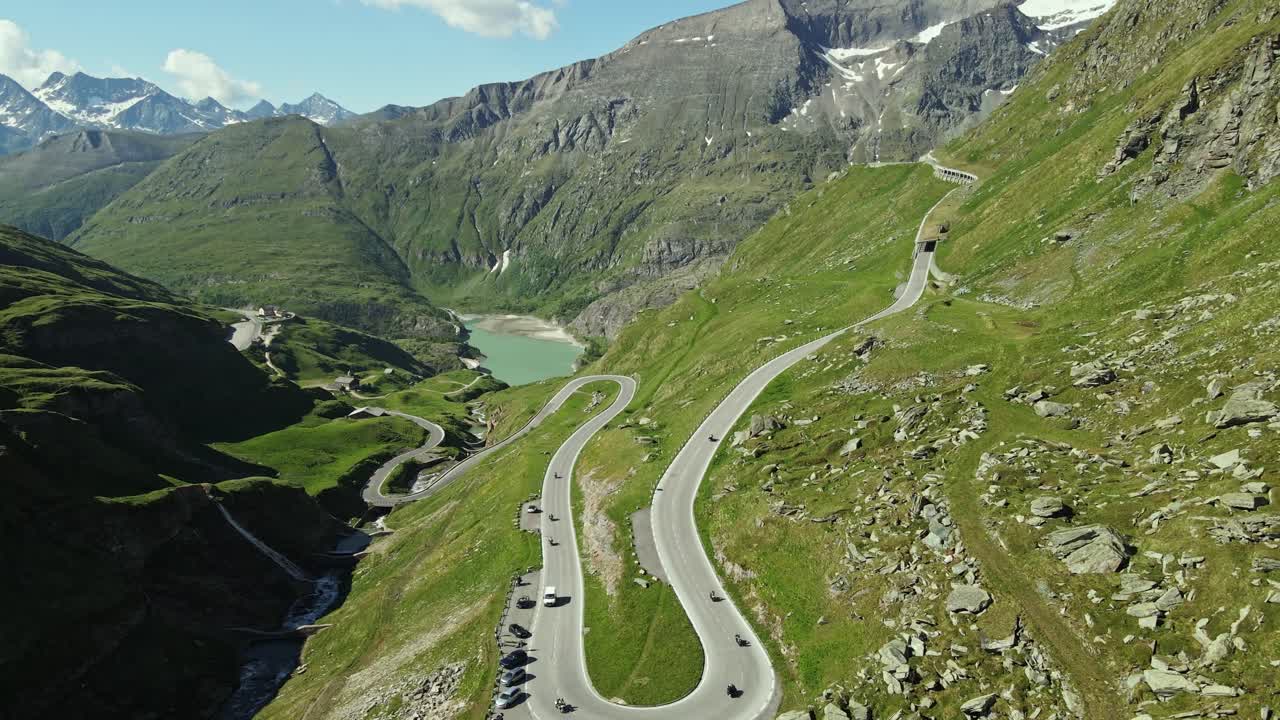 Aerial view of a mountain road filled with motorcyclists and parked cars, surrounded by green alpine slopes. In the background, a turquoise lake nestles among towering peaks under a clear blue sky.