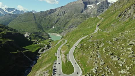 aerial view of a mountain road filled with motorcyclists and parked cars, surrounded by green alpine slopes. in the background, a turquoise lake nestles among towering peaks under a clear blue sky.
