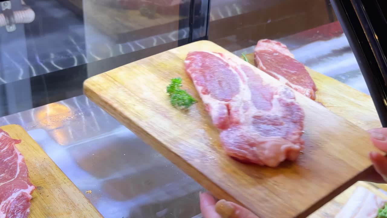 A hand reaches into a display case to select raw pork cuts on wooden boards.