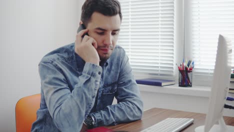 joven trabajador de oficina marcando un número y hablando en su teléfono móvil en la oficina sentado en la mesa con computadora, teléfono y taza. filmado en 4k