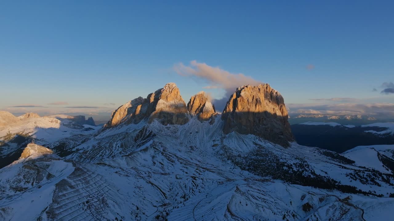 Aerial view of snow-covered peaks glowing in golden sunset light with winding alpine paths across rugged terrain in the Dolomites under clear blue winter sky