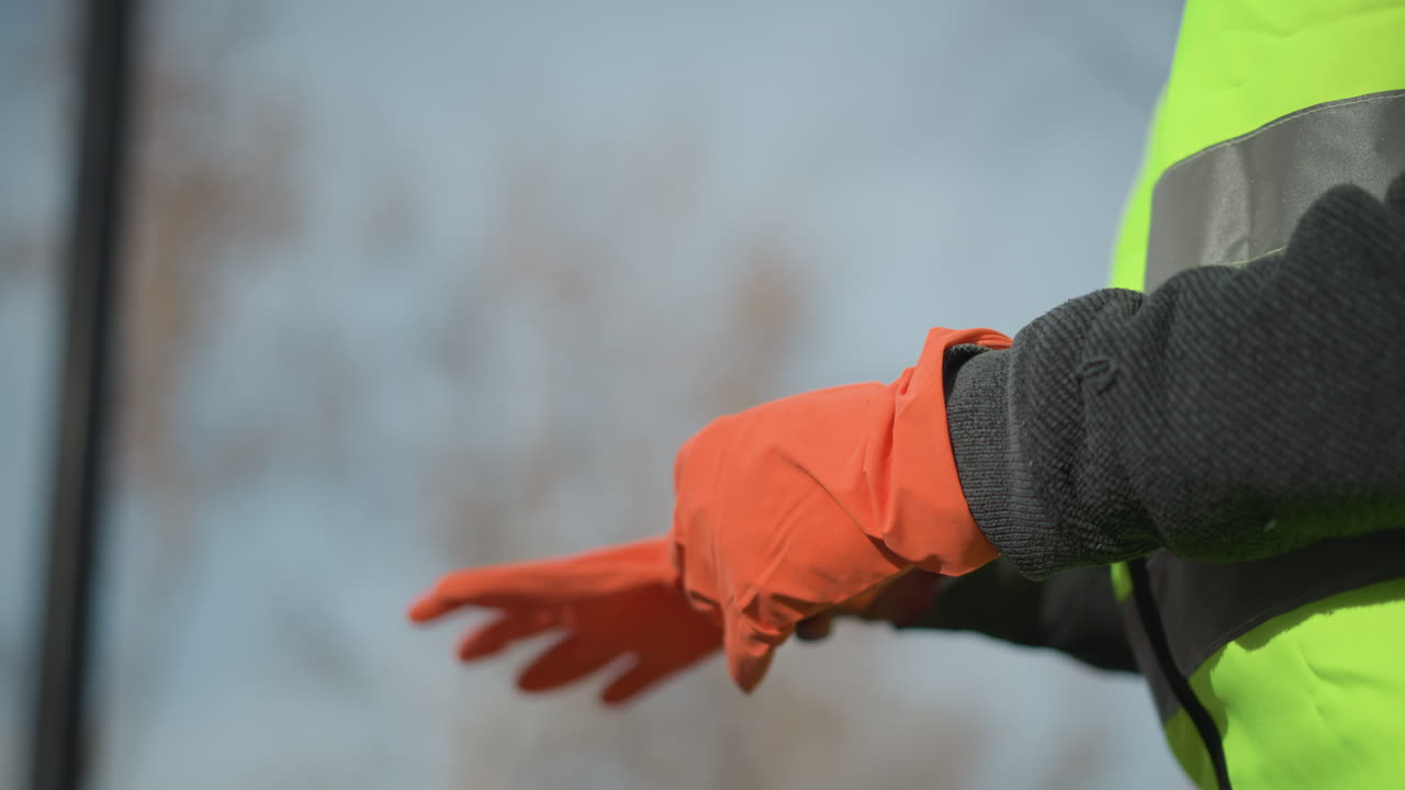 Close-up of worker wearing dark sweater and high visibility vest putting on bright orange protective gloves, preparing for safety tasks in outdoor construction