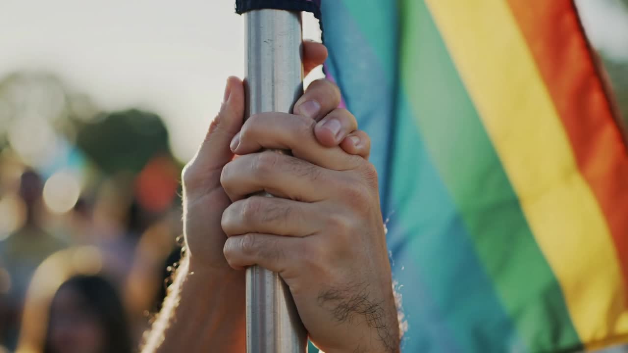 Duas pessoas ajustando o aperto no mastro da bandeira no parque enquanto o vento desenrola a bandeira arco-íris em sinal de apoio