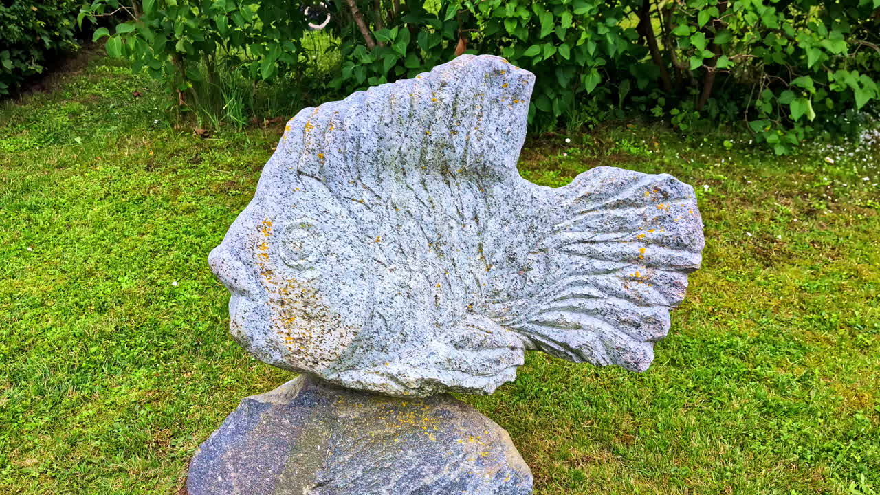 Large stone sculpture of fish placed on grass with green shrub background