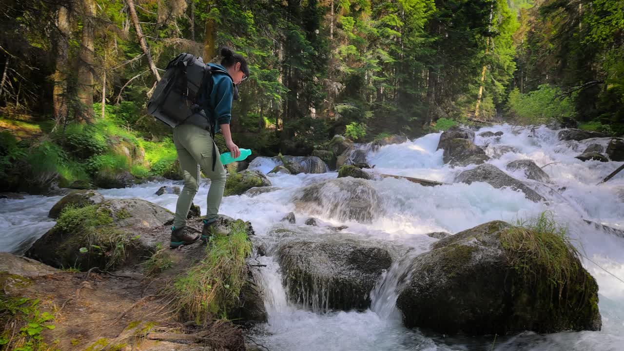 viajera con una mochila, bebiendo agua en la naturaleza en el bosque cerca de un río de montaña.