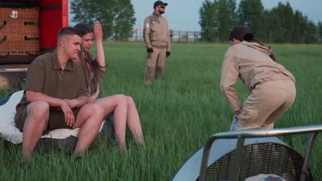 woman in beige flight suit crouching on green grass next to colorful balloon fabric beside fan cage while young couple sits on wicker basket edge by red truck under soft twilight sky