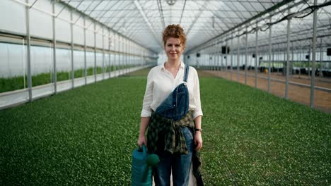 Portrait-of-a-happy-girl-with-curly-hair-in-a-white-farmer-shirt-with-a-watering-can-in-her-hands-on-a-farm-in-a-plant-greenhouse