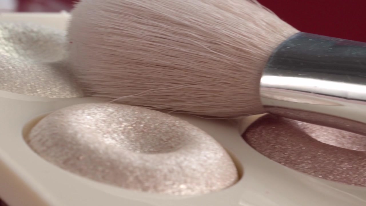 Close up panning shot of makeup brush gliding across shimmering highlighter palette showcasing cosmetic powder texture beauty products and makeup application
