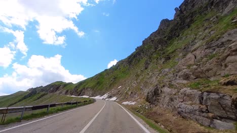 Slow-Drive-Up-A-Mountain-Road,-Driver-POV,-Tall-Mountain-Side-With-Patches-Of-Snow,-Clear-Blue-Sky,-Transfagarasan-Highway,-Romania