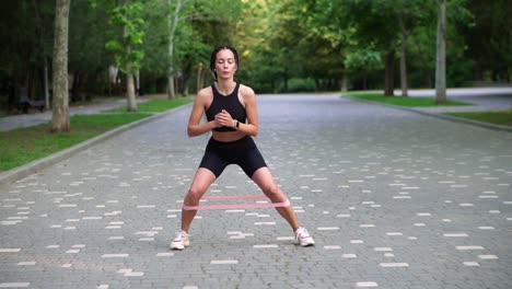 Front-view-of-stylish-woman-makes-crunchy-side-steps-with-pink-rubber-band-on-legs-on-pavement,-view-on-green-city-park-on