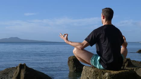 Junger-Fitter-Mann,-Der-Morgens-In-Yoga-pose-Auf-Felsen-Am-Strand-Meditiert