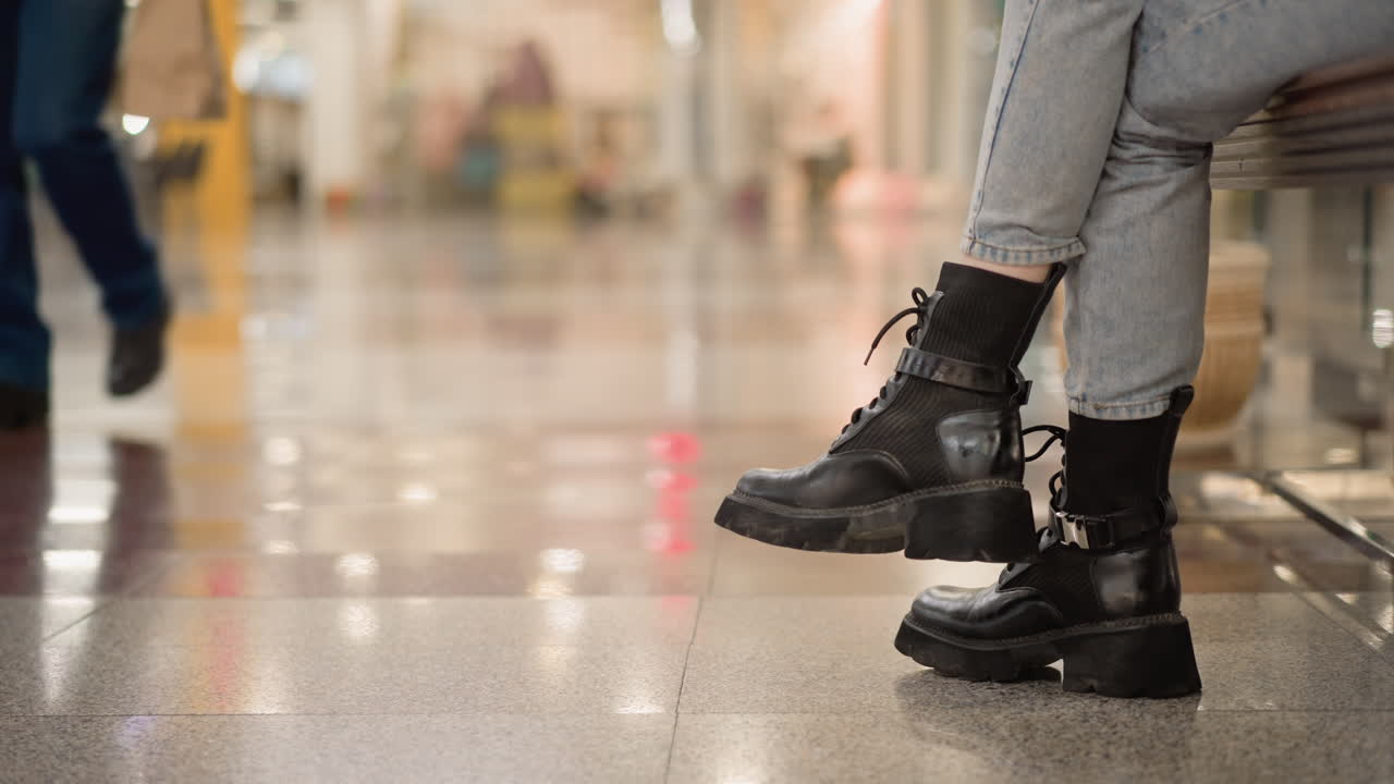crossed leg of shopper seated on mall bench wearing chunky black boots and denim jeans with blurred male leg walking past under bright shopping mall ceiling lights reflecting on polished floor