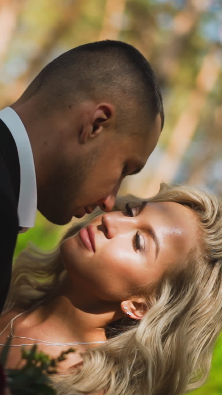 Careful gentleman in classic suit kisses cheek of beloved blonde wife with hairstyle closing eyes standing in sunny spring forest close view slow motion