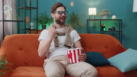 young man sits on sofa eating popcorn and watching interesting tv serial, sport game online at home