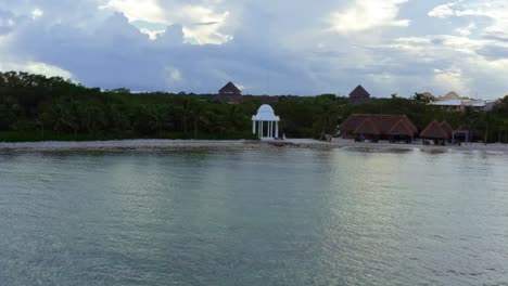 Beautiful-dolly-out-aerial-drone-shot-of-the-tropical-coastline-of-playa-del-carmen-with-large-vacation-resorts-and-a-small-white-gazebo-in-Riviera-Maya,-Mexico-on-a-warm-sunny-summer-day